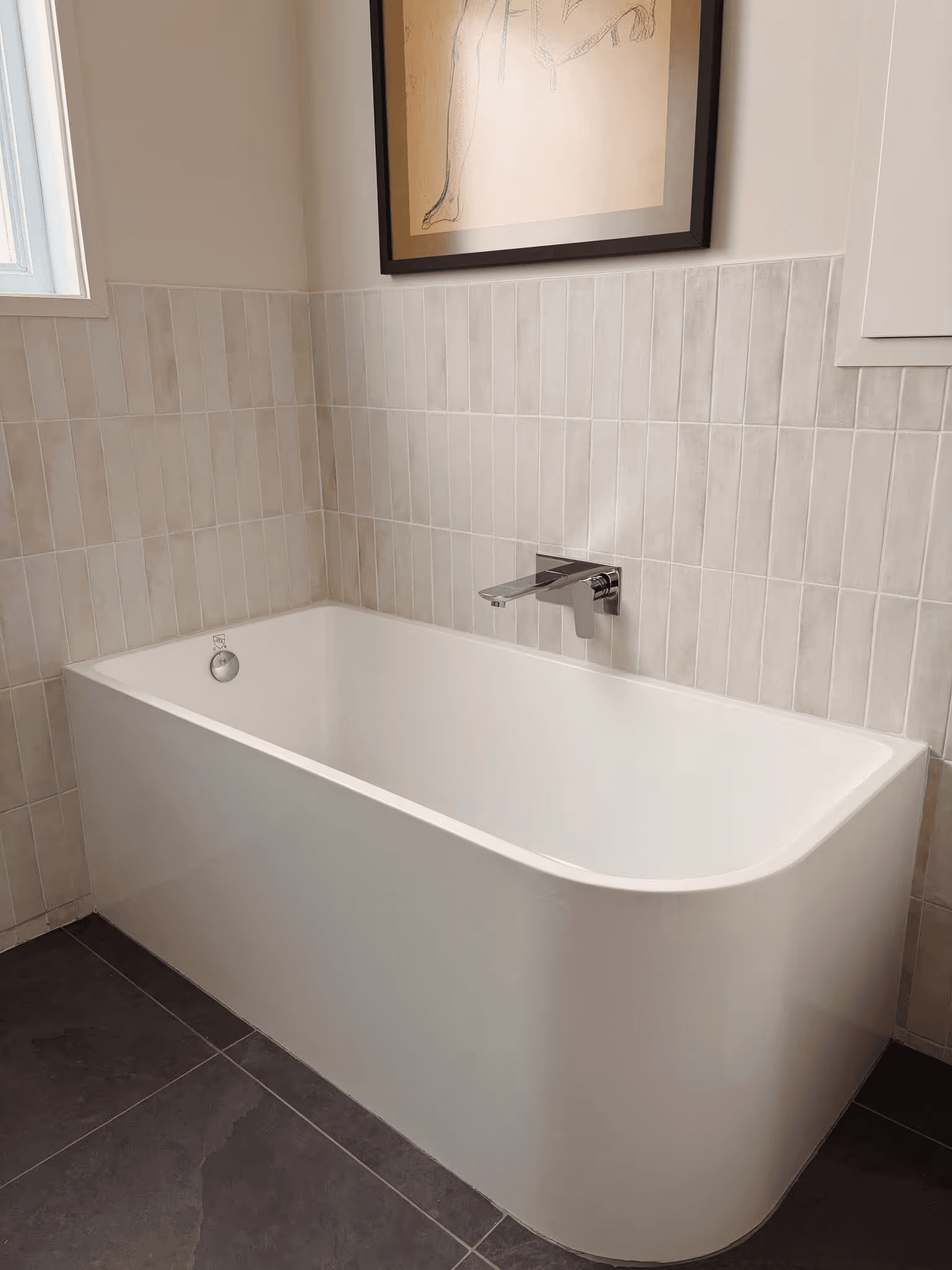 Modern bathroom featuring a white freestanding rectangular bathtub with rounded corners, chrome wall-mounted faucet, vertical beige wall tiles, dark gray ceramic floor tiles and minimalist decor