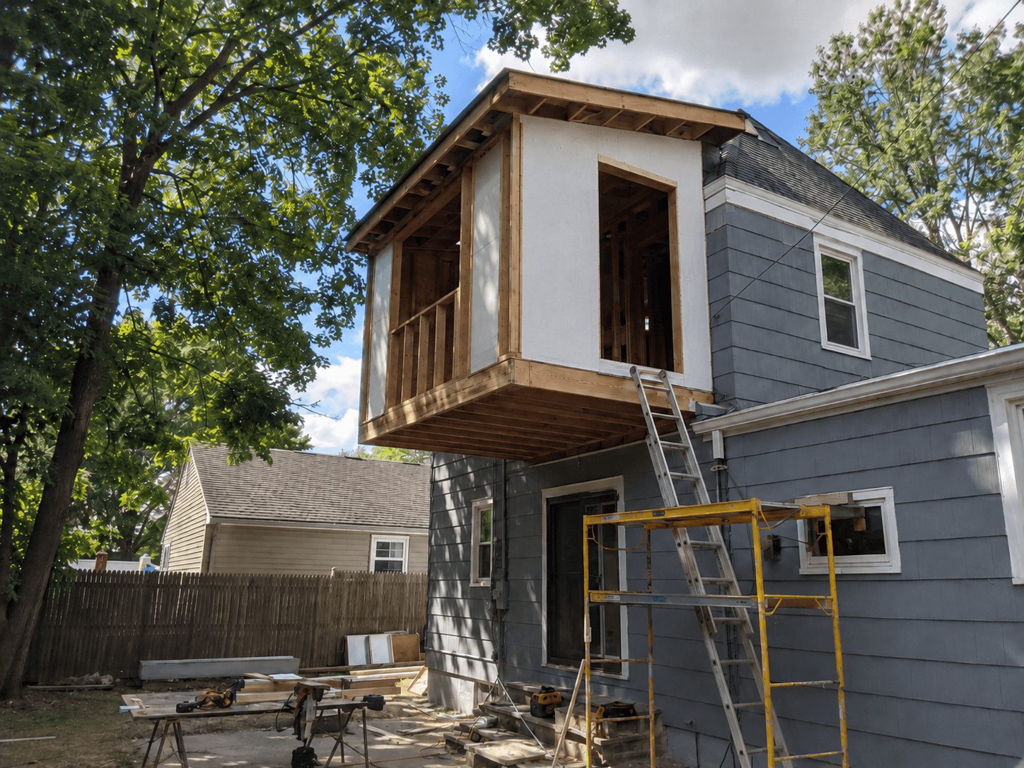 Cantilevered second-story rear home addition under construction with wood framing, structural sheathing, large openings, scaffolding, and a ladder