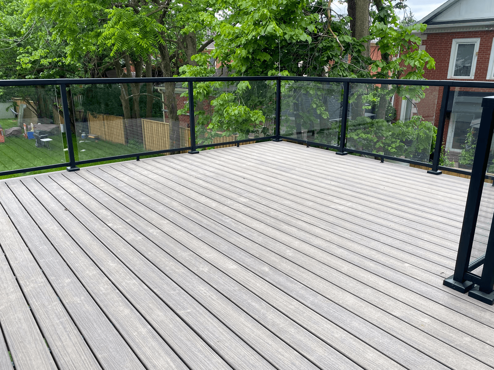Large elevated residential deck with grey wood decking, modern transparent glass railings and a black aluminum structure overlooking a green backyard with mature trees and brick houses.
