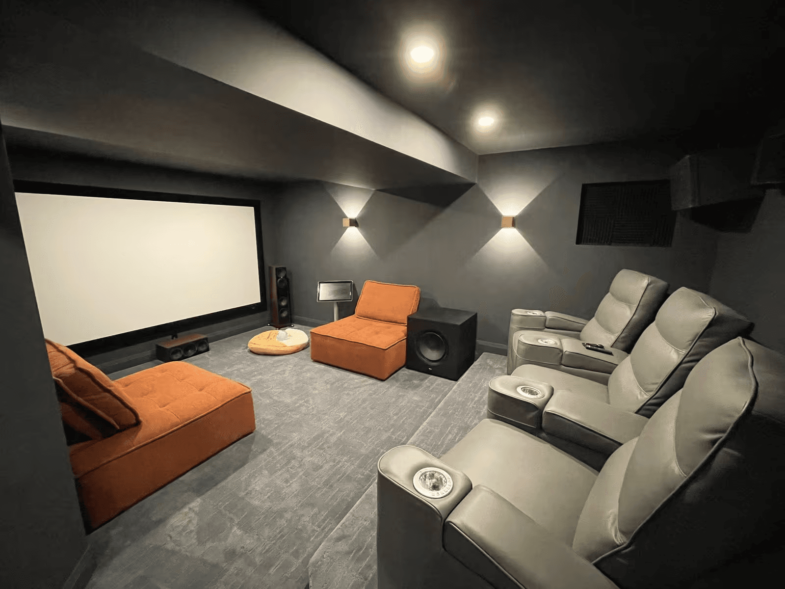 Dark home theatre with large projection screen, grey reclining chairs, orange lounge seating, surround sound system and modern wall lighting