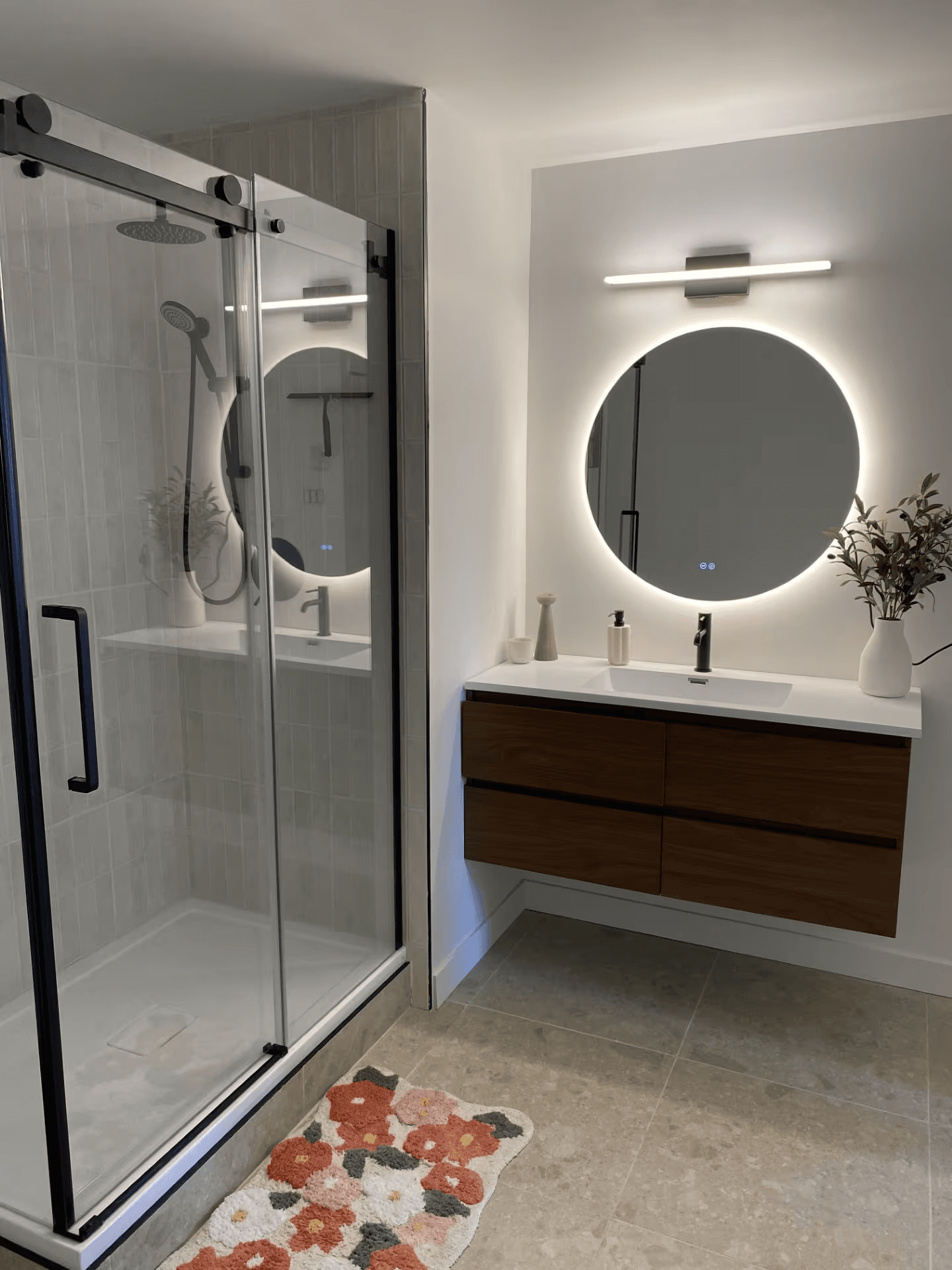 Modern renovated bathroom with a glass-enclosed shower, black fixtures, floating wood vanity and illuminated round mirror.