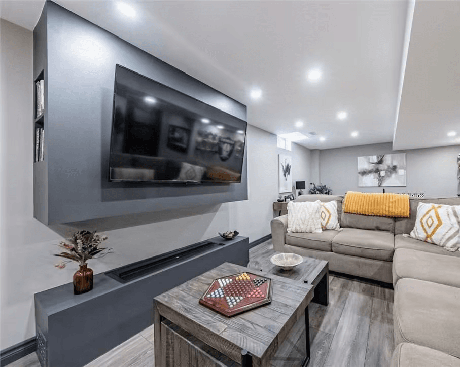 Modern basement family room with large wall-mounted television, grey sectional sofa, wood tables, recessed lighting and wood-look flooring