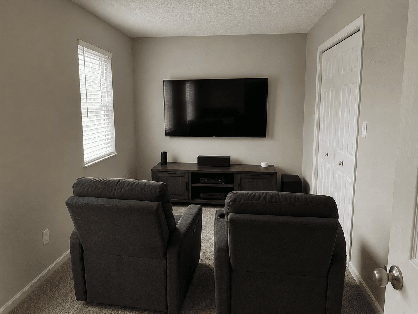Small home theatre with a wall-mounted TV, media console, audio system, two reclining chairs and carpet in a residential room.