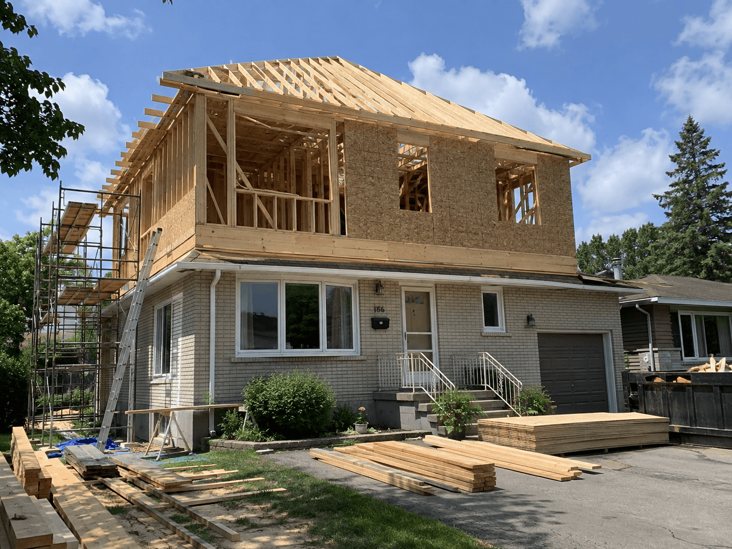 Residential second-story addition under construction with wood framing, roof trusses, structural sheathing, scaffolding, and building materials