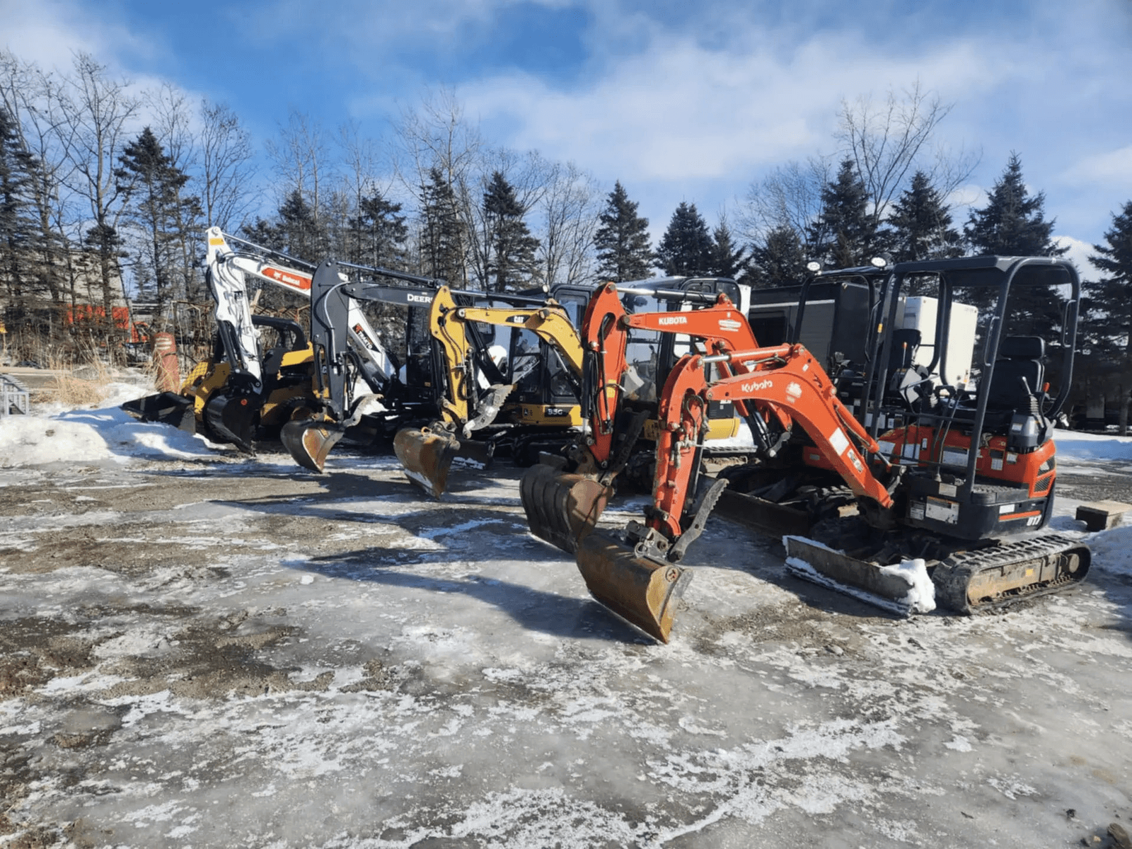 Lineup of compact excavators and tracked construction equipment parked on a snowy lot, including Kubota, Caterpillar, Bobcat and John Deere machines in front of a wooded area.