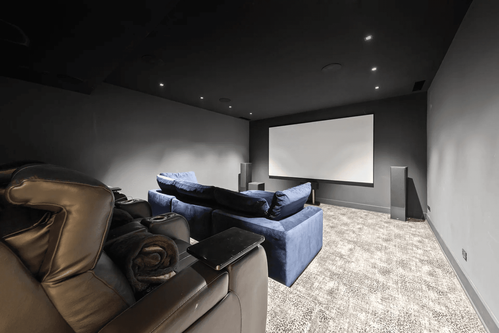 Dedicated home theatre with a large projection screen, speakers, reclining chairs, sofa and recessed lighting in a dark room.