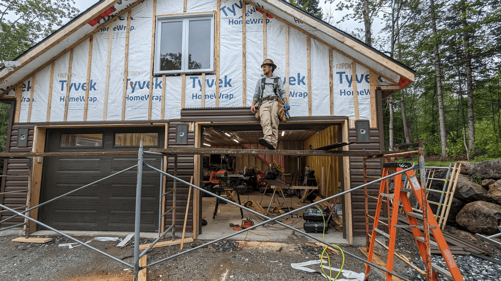 Construction worker in front of a house under renovation with weather barrier, scaffolding, ladders, tools and partially installed exterior siding