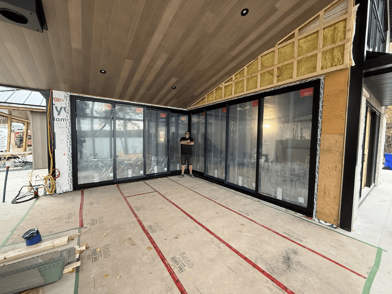 Large residential extension under construction with floor-to-ceiling black sliding glass doors, finished wood ceiling, exposed insulation and panoramic outdoor opening.