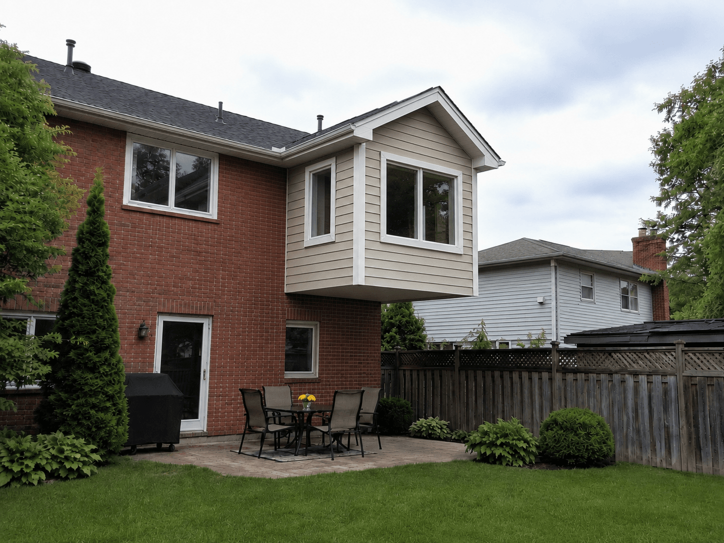 Cantilevered rear home addition on a brick house with large windows, beige siding, gabled roof, and landscaped backyard patio