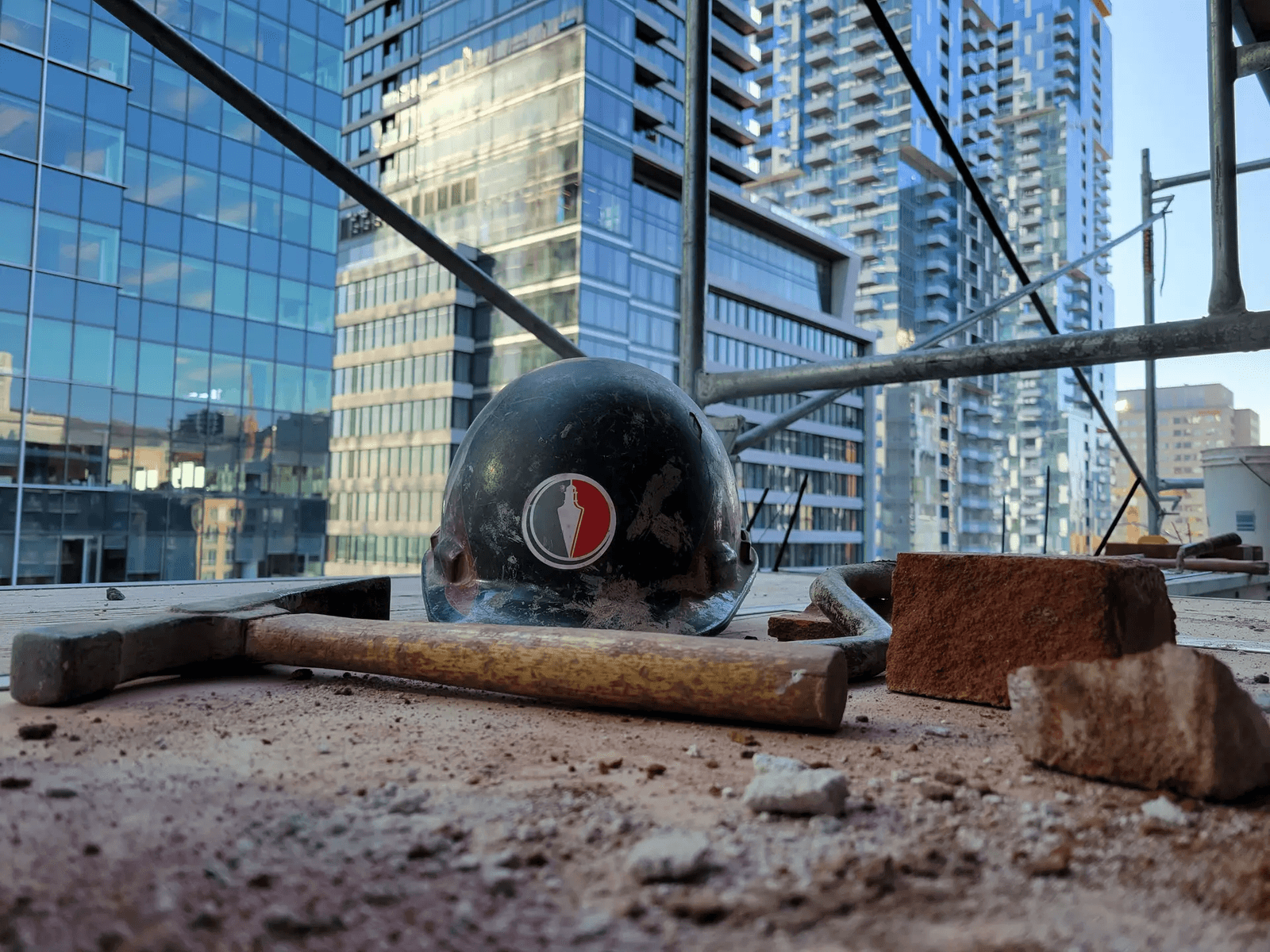 View of a high-rise construction site with a safety helmet, hammer, and bricks, surrounded by modern buildings in the background.