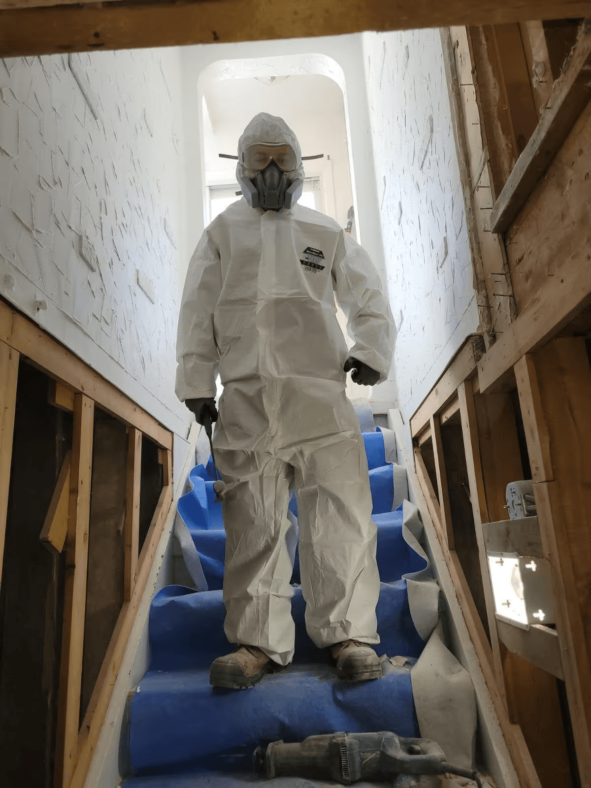 Worker wearing a protective suit and respirator on a staircase under renovation, surrounded by open walls and exposed framing
