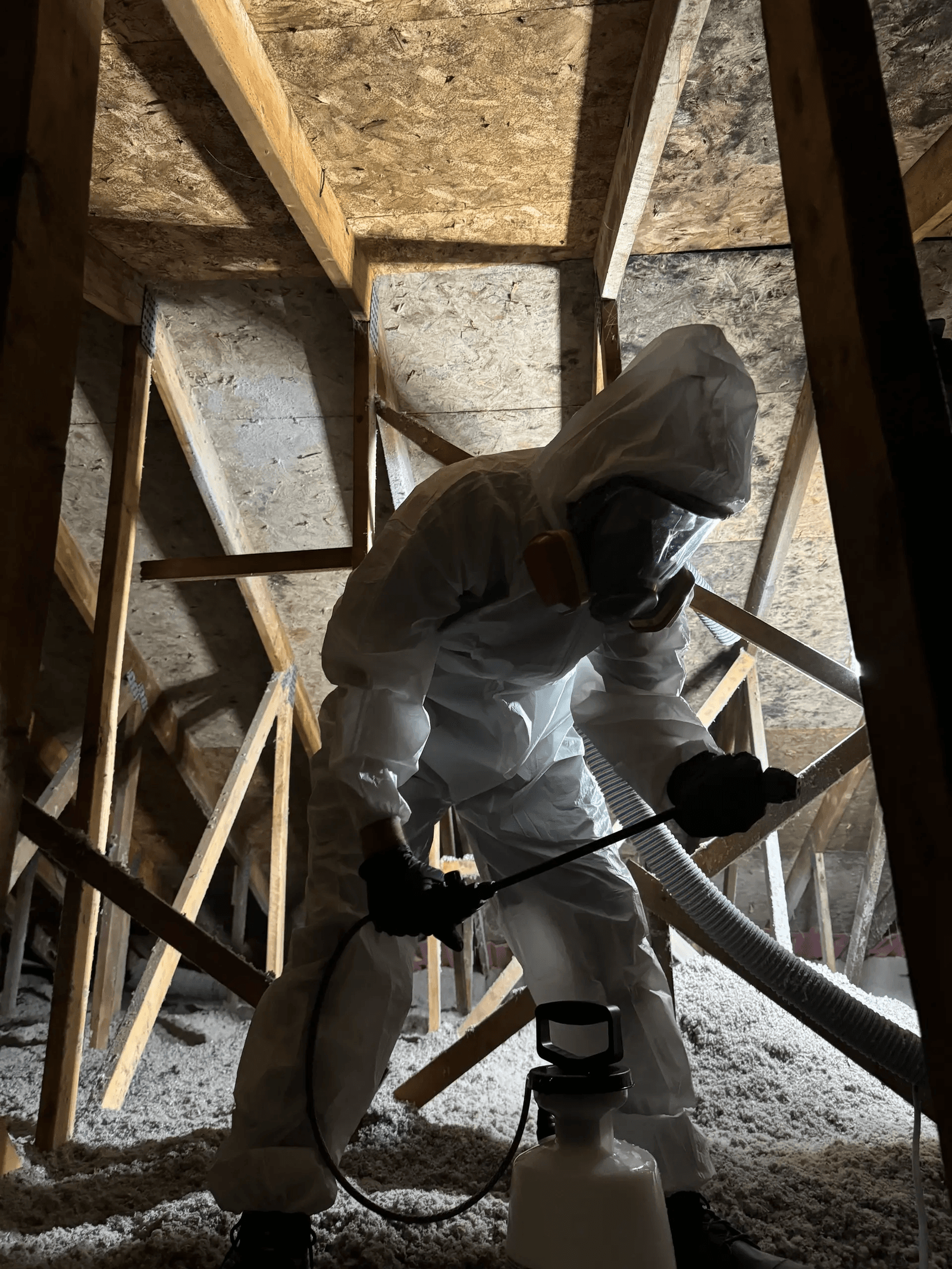 Technician wearing white protective coveralls, a filtered respirator and black gloves using a handheld pressure sprayer inside a residential attic with wooden roof framing and blown-in insulation, illustrating specialized attic treatment and decontamination work