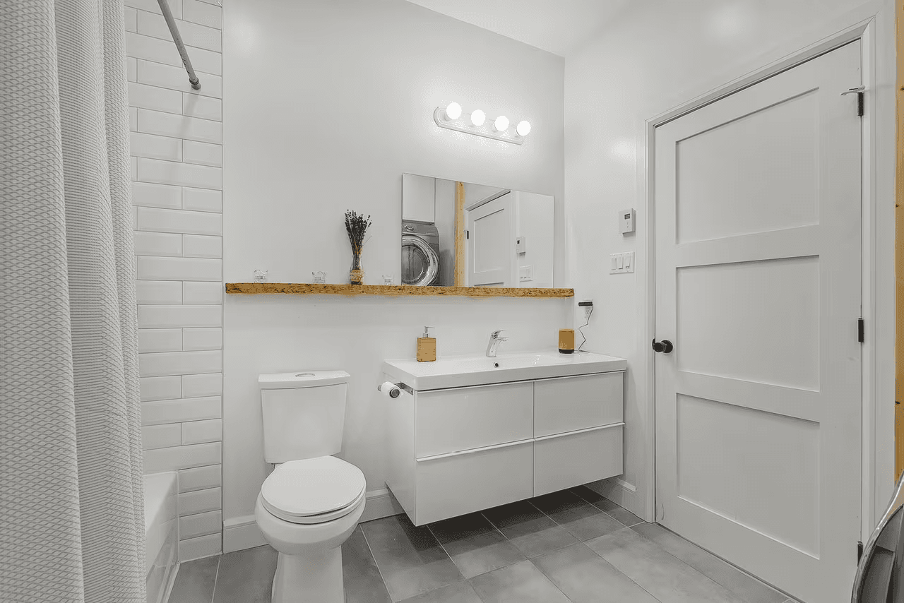 Clean modern bathroom with a floating white vanity, wall mirror, toilet, tiled shower, gray flooring, and a white interior door