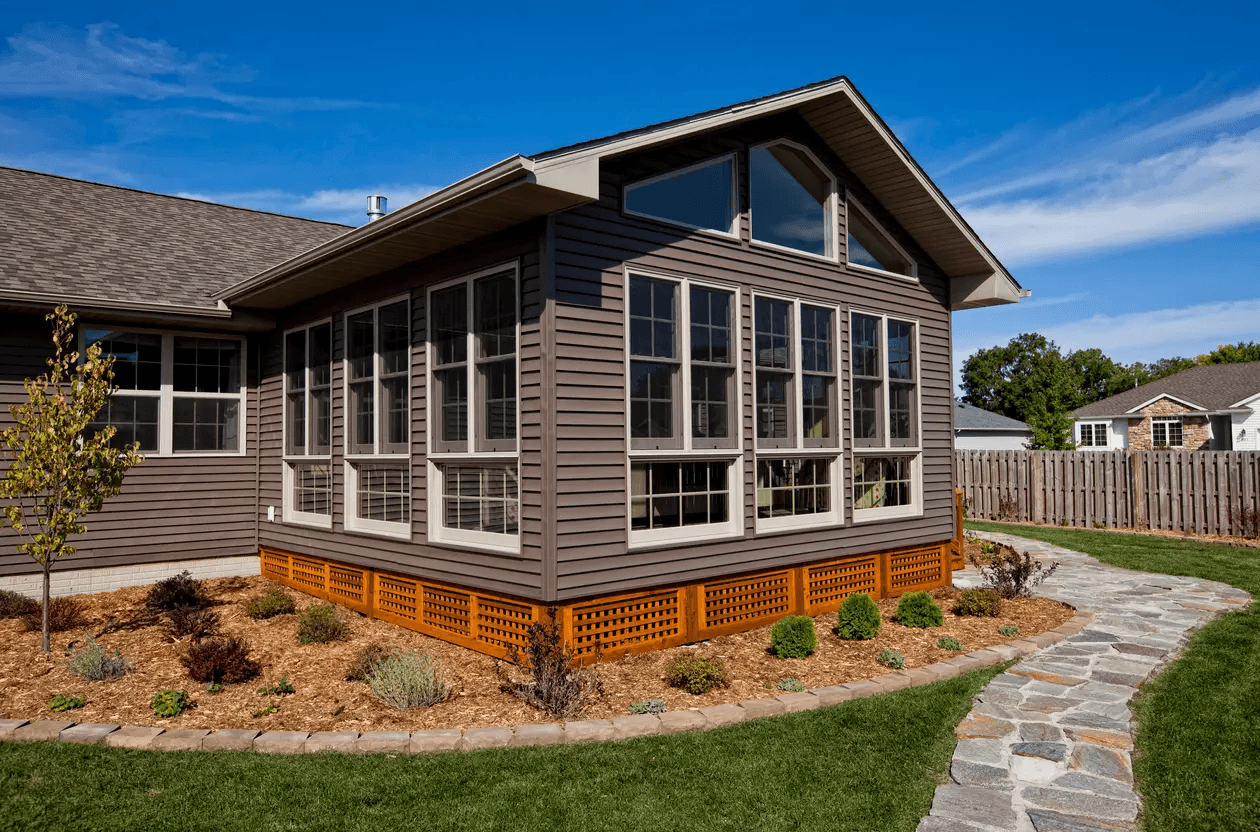 Home addition with a large glass veranda, wood siding, landscaped garden with mulch and a stone walkway, and a blue sky in the background.