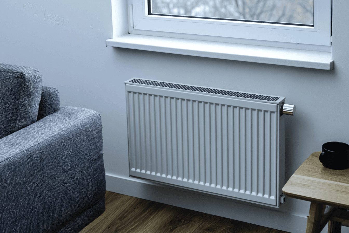 White panel radiator mounted beneath a window in a modern living room with grey sofa and hardwood flooring