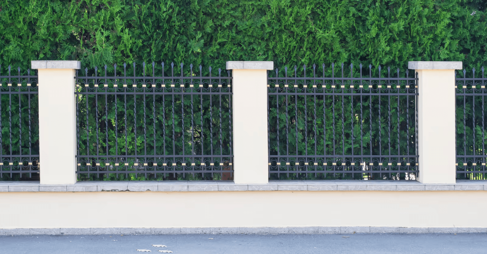 Decorative black metal fence with pointed pickets and circular accents, installed between cream-colored masonry pillars on a low wall, with a dense green evergreen hedge in the background.