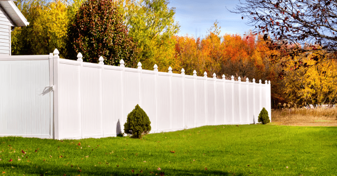 White vinyl privacy fence with decorative post caps enclosing a green residential lawn, surrounded by colorful autumn trees and landscaping.