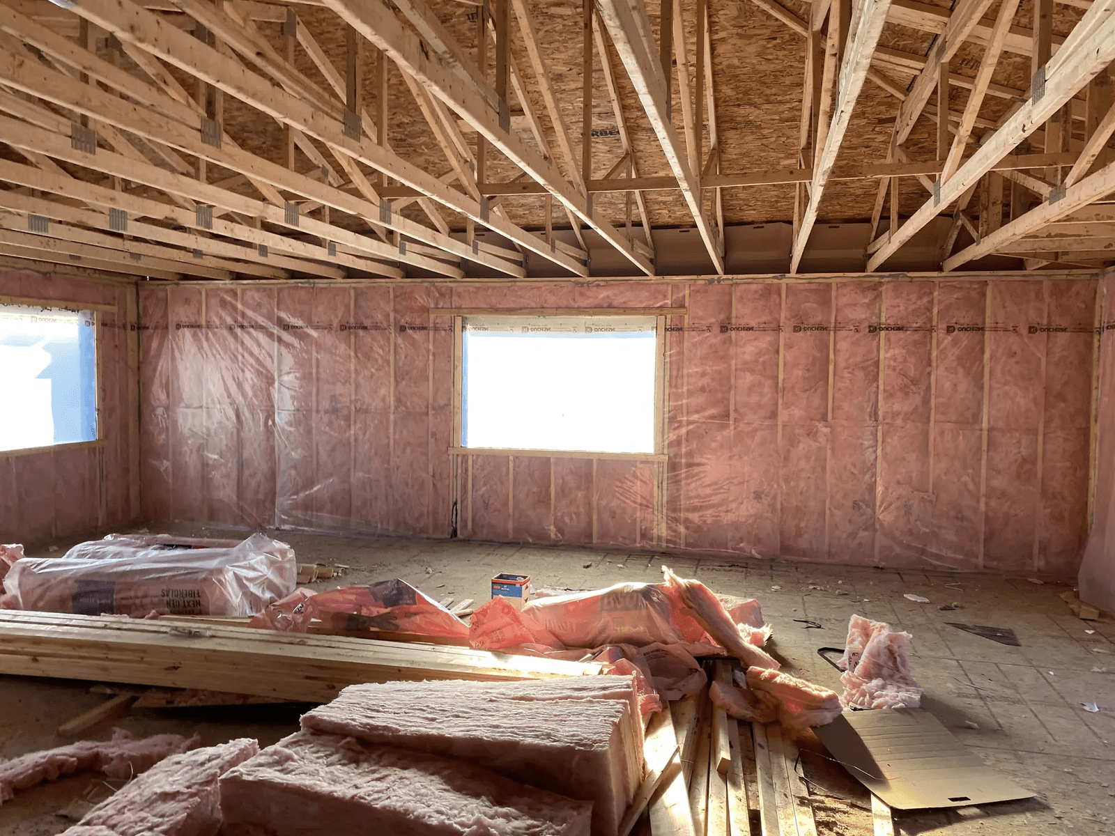 Residential interior under construction with exposed roof framing, walls insulated with pink fiberglass, vapor barrier, and construction materials on the floor.