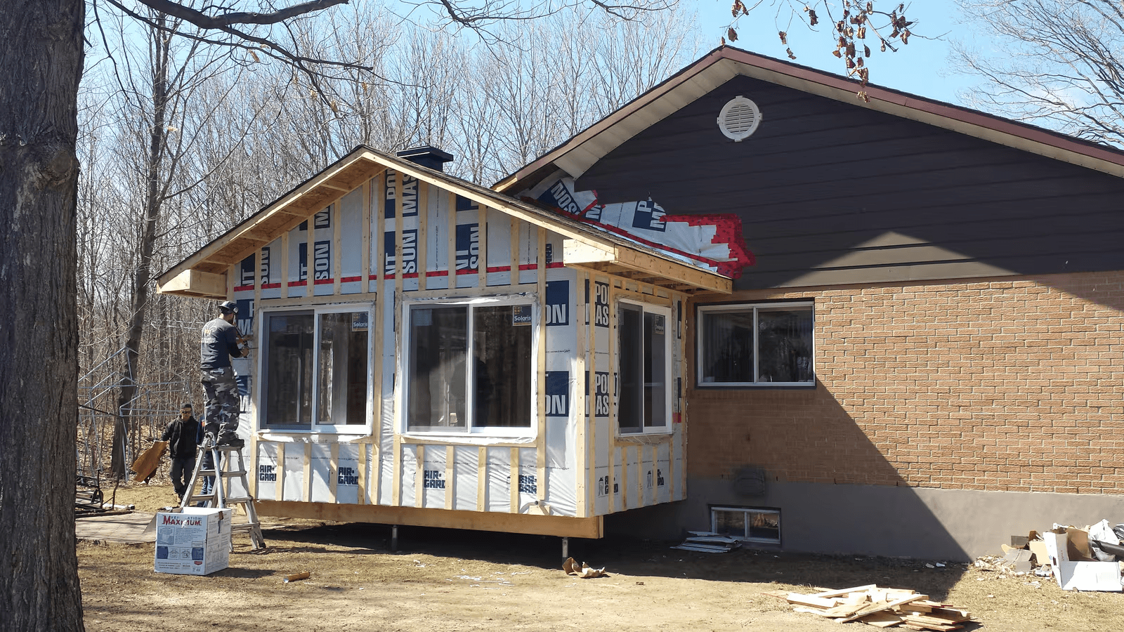 Home addition under construction with wood framing, large windows, weather-resistant membrane, and exterior siding installation.