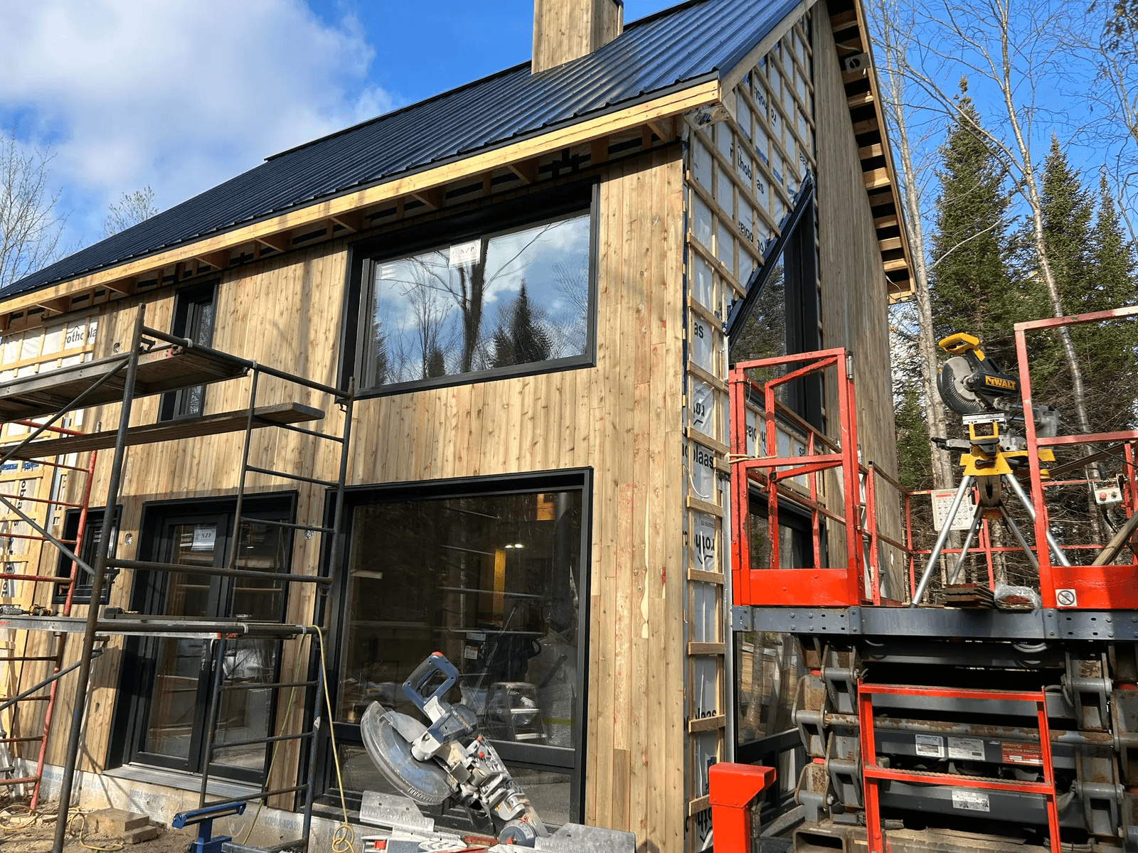 House under construction with wooden exterior cladding, large windows, and a black metal roof, surrounded by scaffolding and tools.