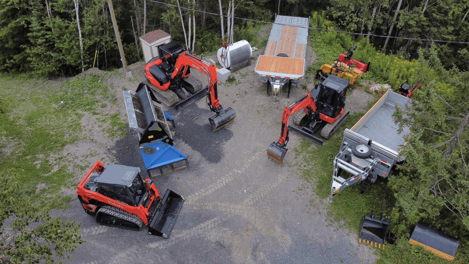 Aerial view of a heavy equipment yard with compact excavators, tracked loader, trailers and excavation attachments on a gravel lot surrounded by forest.