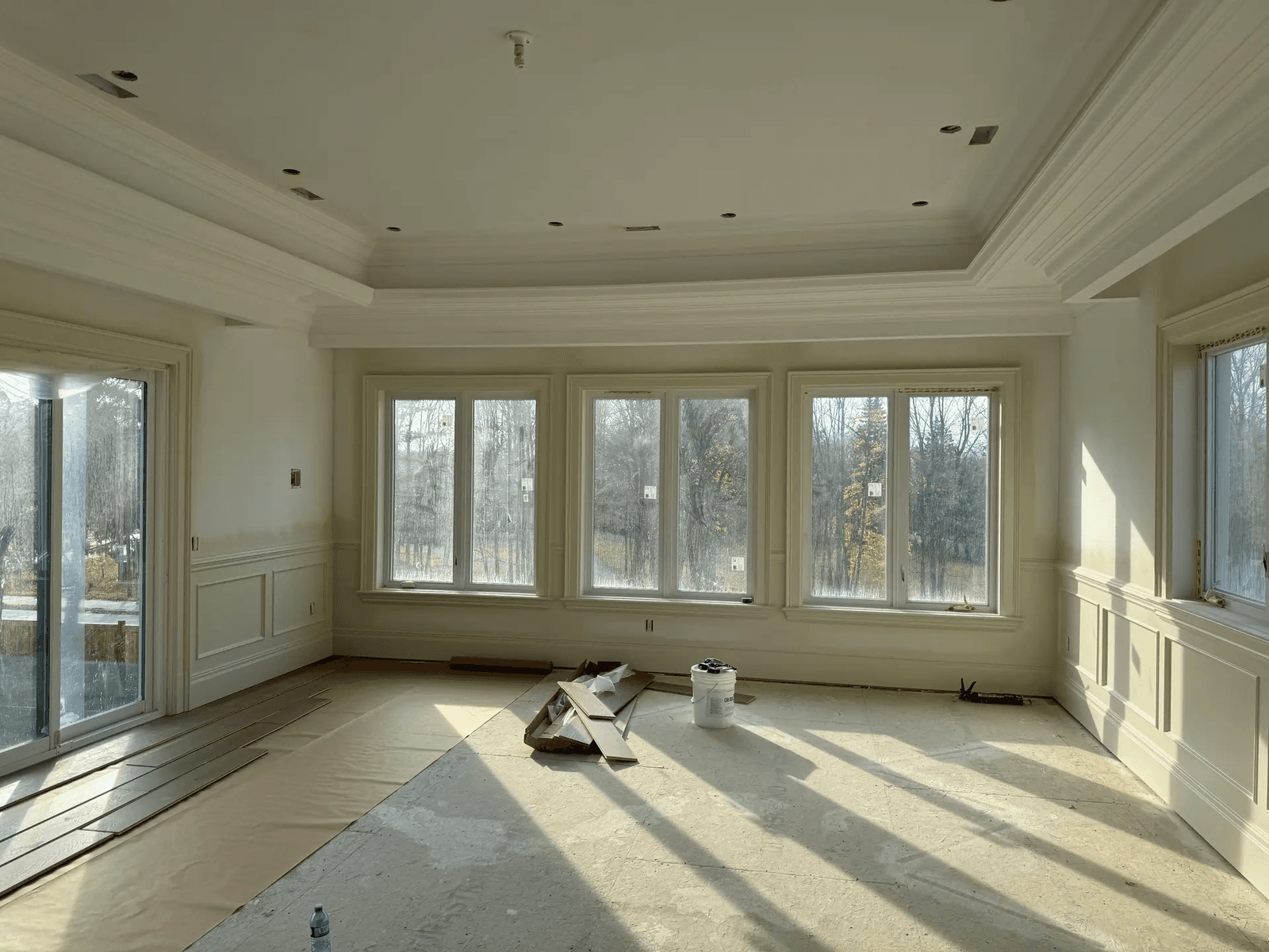 Bright room under renovation with decorative moldings, large windows, coffered ceiling, and flooring under installation.