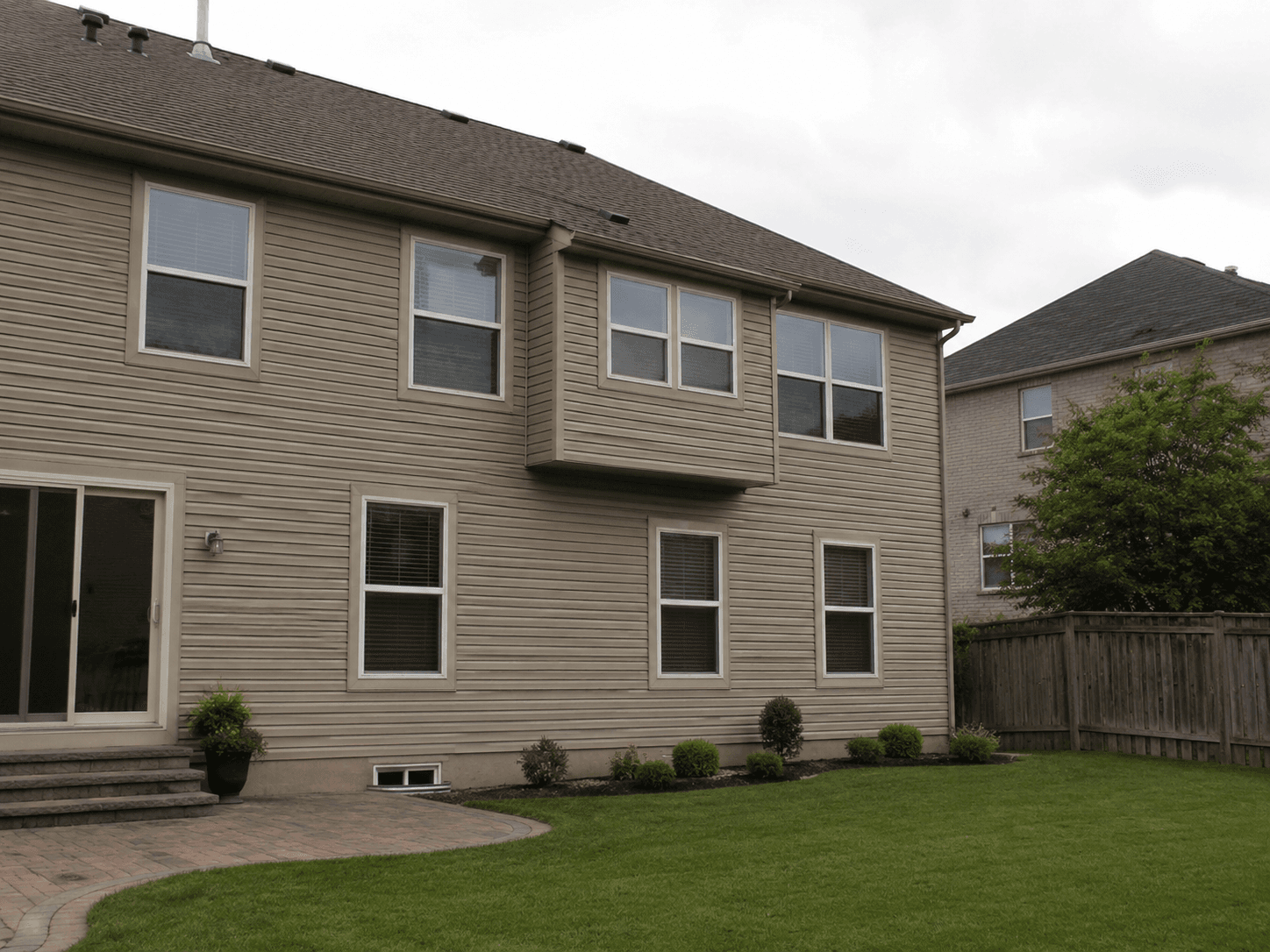 Rear view of a residential house with beige exterior siding, multiple windows, shingle roofing, a small cantilevered projection, and a fenced grassy backyard