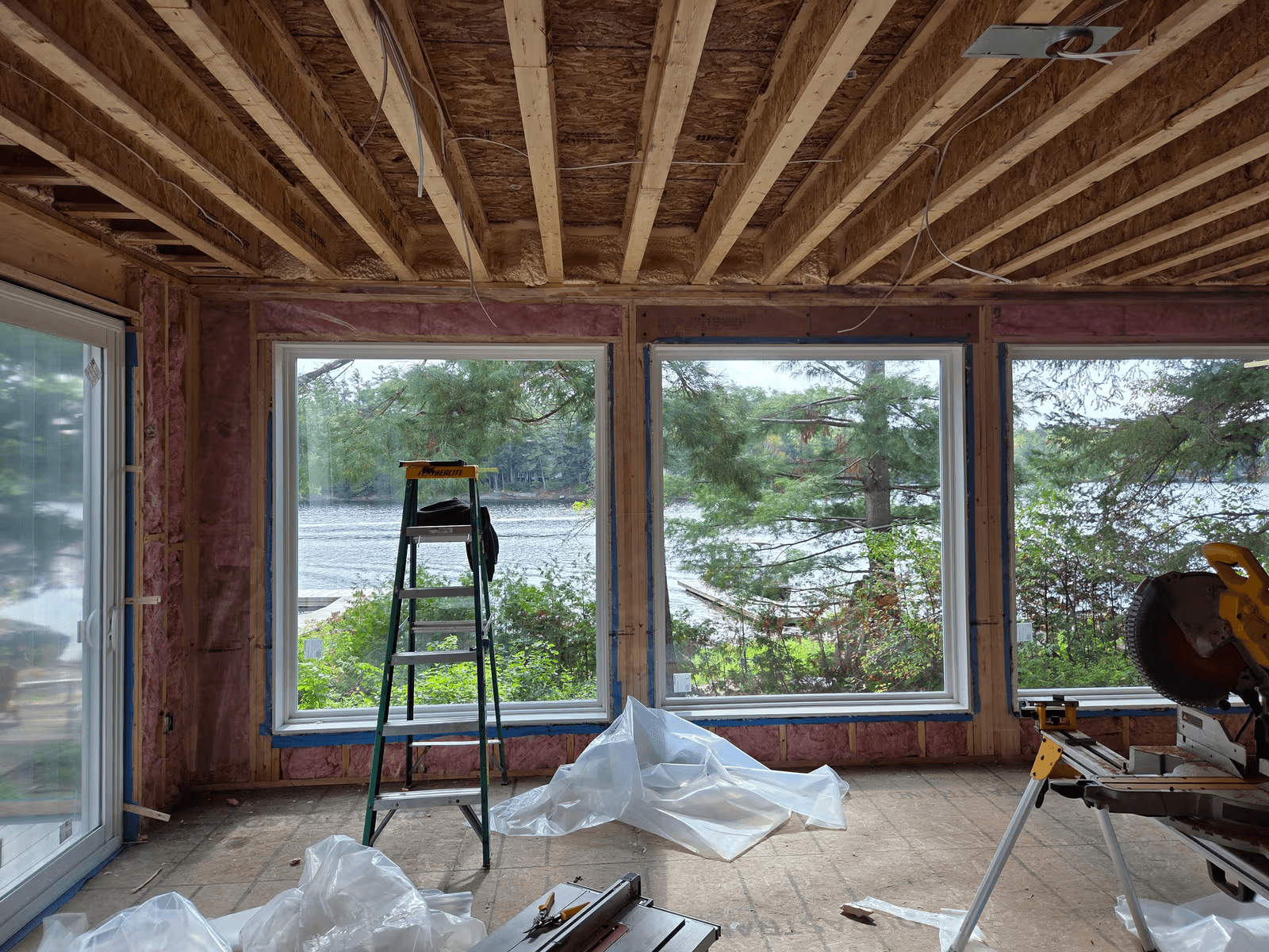 Residential room under renovation with exposed framing, fiberglass insulation, large windows overlooking the water, and construction tools.