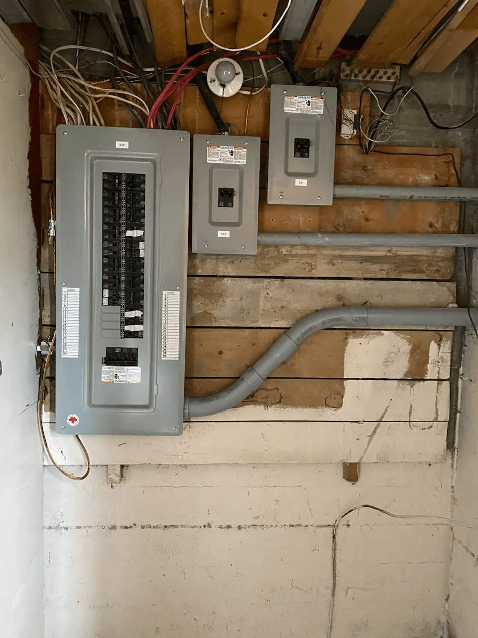 Main electrical panel and subpanels installed on an unfinished wooden wall with gray conduits and exposed wiring