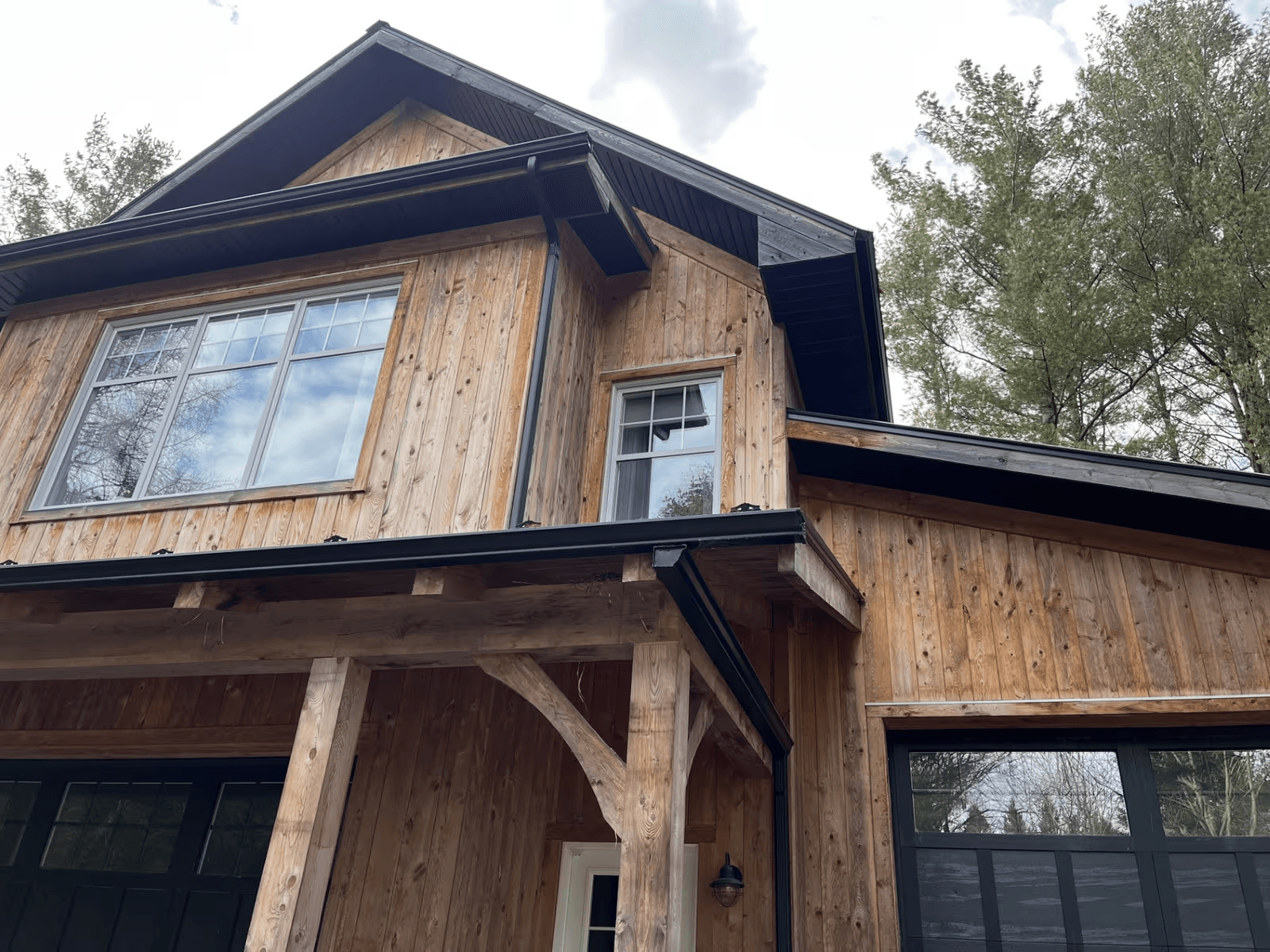Exterior facade of a two-story residential house featuring natural vertical wood siding, dark roof shingles, black soffits and fascia boards, black gutters, large white-framed windows, black garage doors and a covered entrance porch with solid timber beams and posts