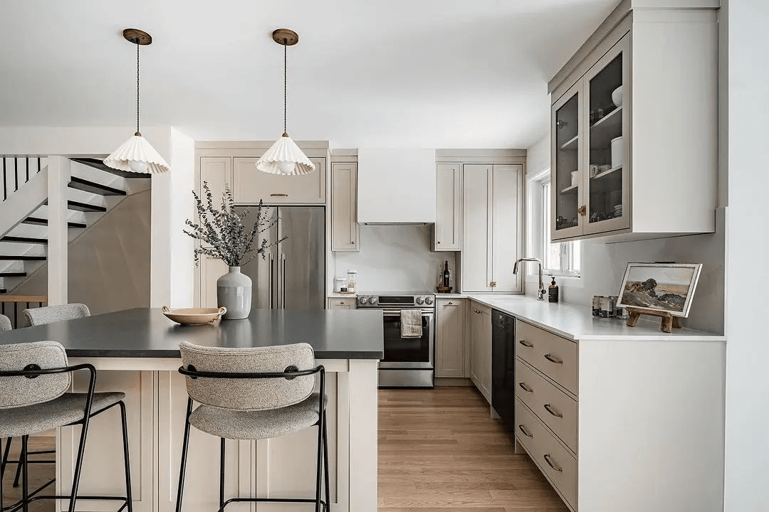 Transitional-style kitchen in Montreal featuring a central island with a dark quartz countertop. Beige cabinets and brass handles add elegance, while light wood flooring and upholstered bar stools create a warm and inviting atmosphere.