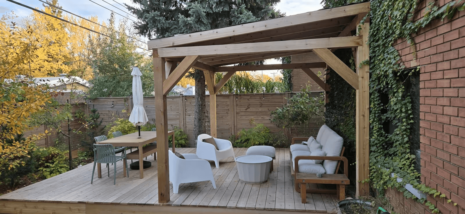 Residential natural wood deck with a sloped-roof pergola, exposed beams, sturdy posts, an outdoor living area with white sofas and armchairs, a dining table, wooden privacy fencing and lush landscaping beside a brick house.