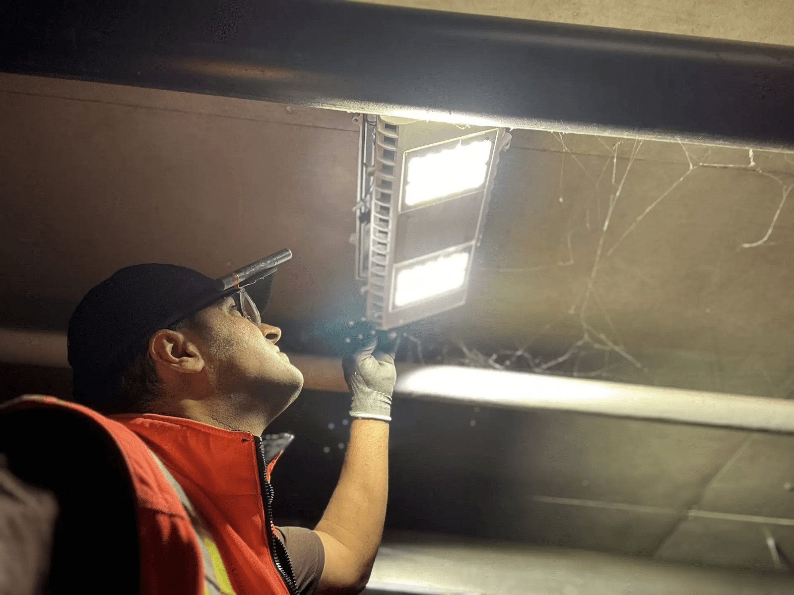 Lighting technician inspecting or adjusting an LED fixture installed on the ceiling in a dark indoor space.