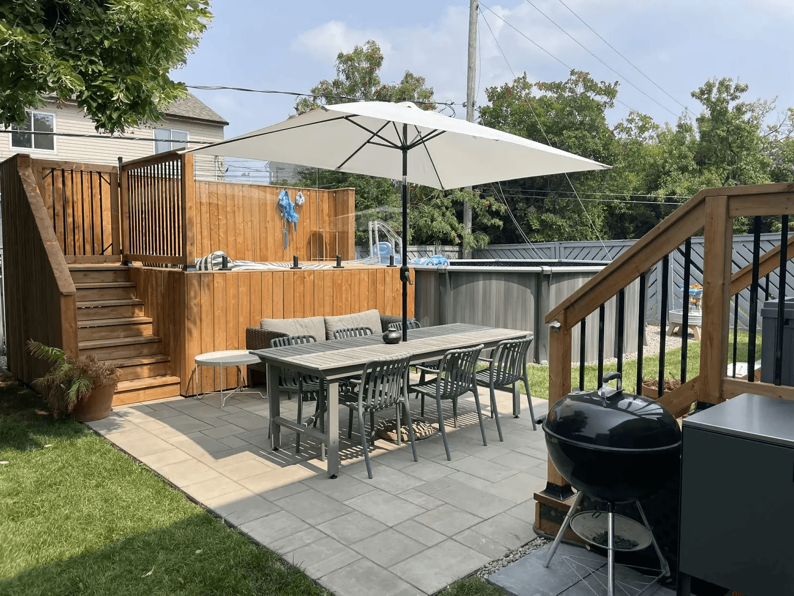 Residential backyard featuring a grey stone patio with an outdoor table beneath a large white umbrella, an elevated wooden deck with stairs and black railings, an above-ground pool, privacy fencing and a barbecue.