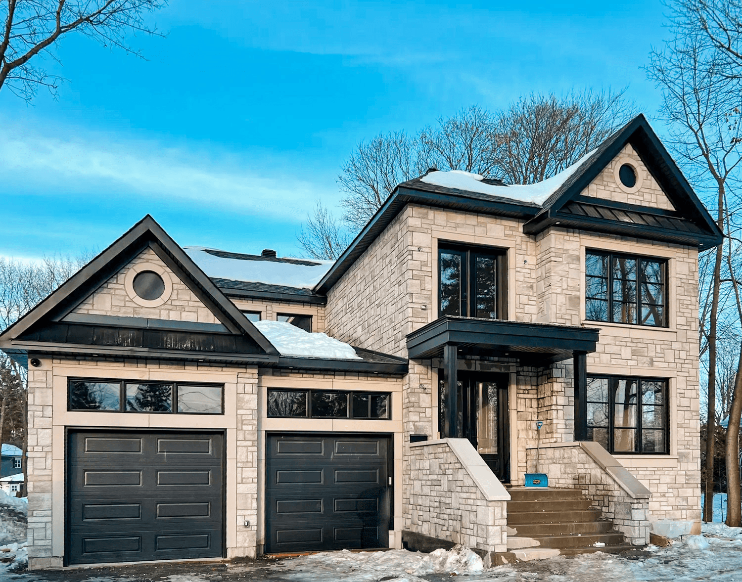 High-end stone house with two black garages, large windows, and snow-covered roof in winter.