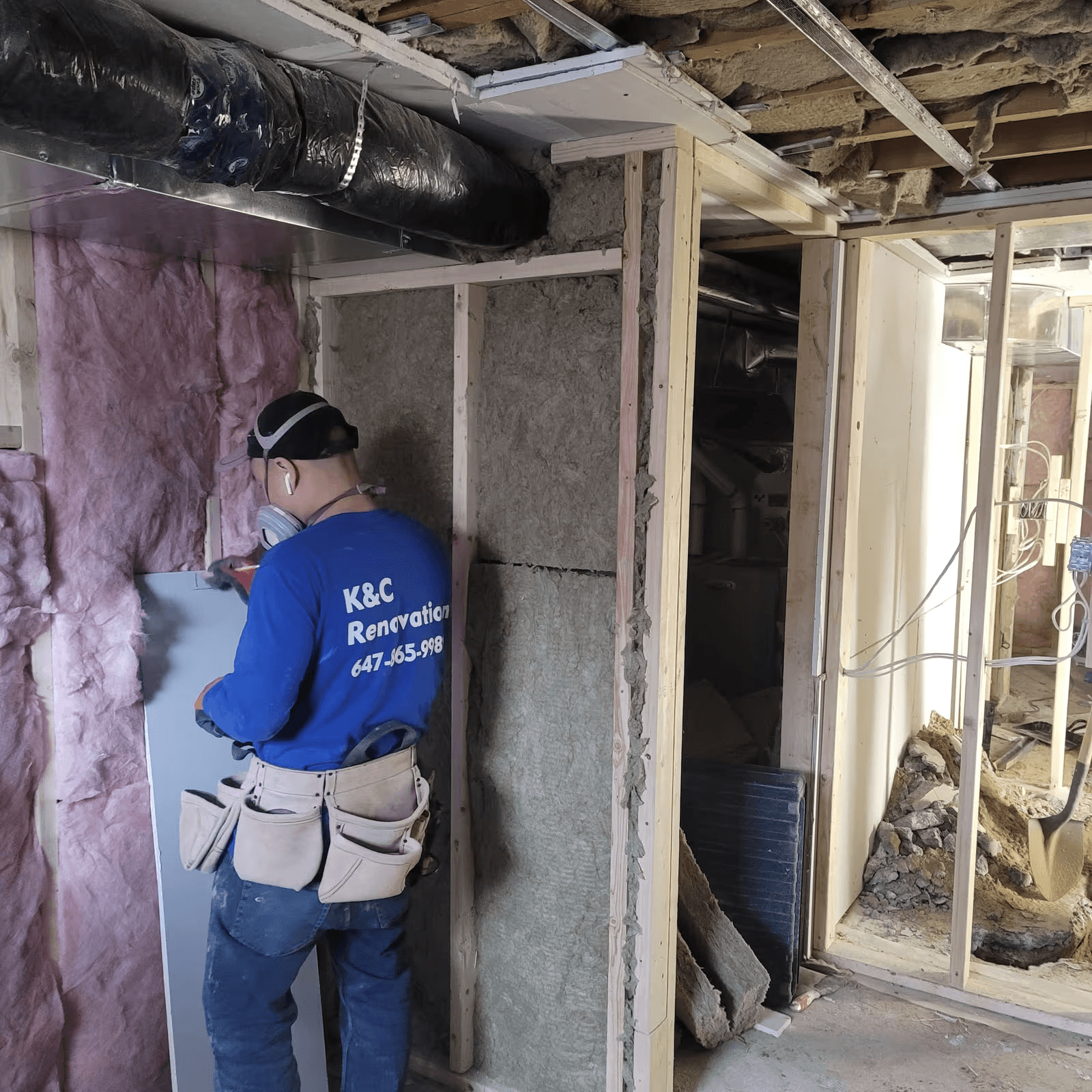 Renovation worker installing drywall panels in a room under construction with wood framing, fiberglass insulation, and exposed ventilation ducts.