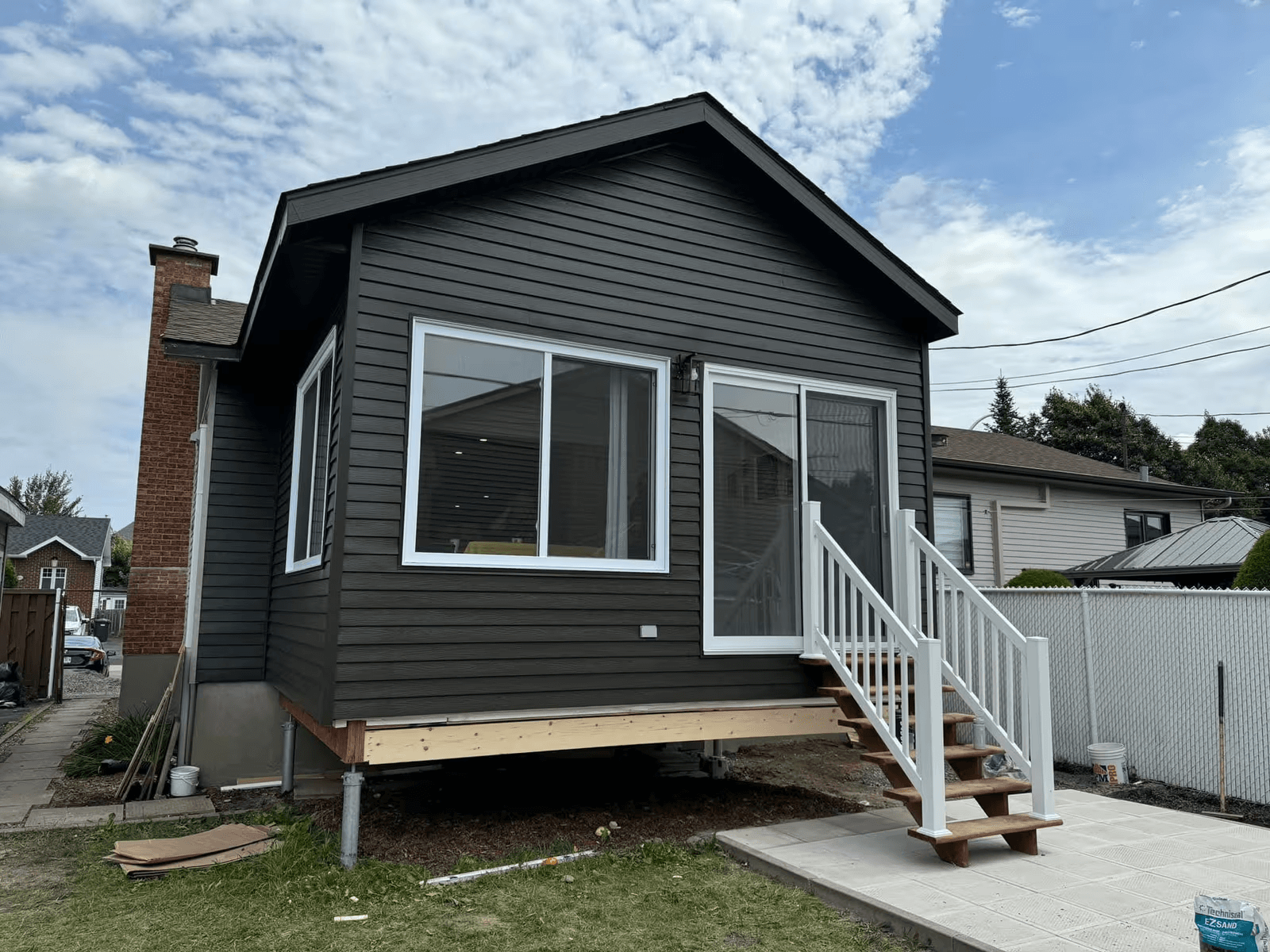 Home addition supported by metal screw piles, featuring charcoal-grey exterior siding, large windows, a patio door, and an exterior wood staircase with white railings.