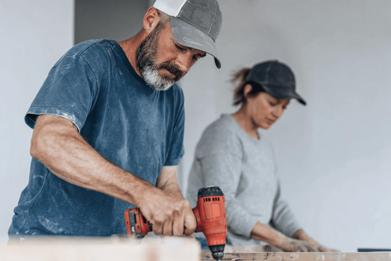Interior renovation workers using a power drill and hand tools on a work surface inside a room under construction.