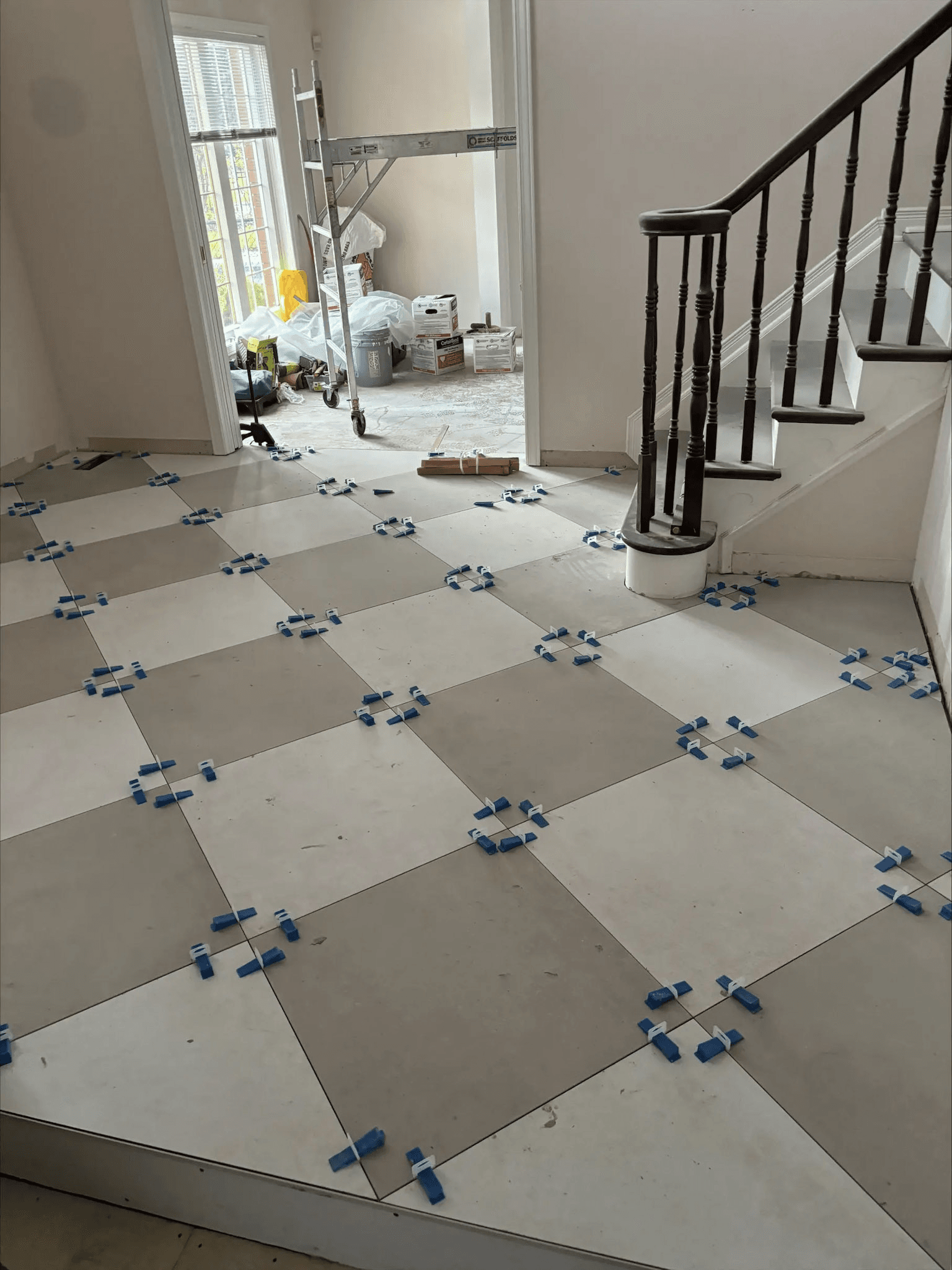 Installation of large-format gray and white ceramic floor tiles in a checkerboard pattern in a residential entryway under renovation, featuring blue tile leveling spacers, a dark wood staircase and construction materials.