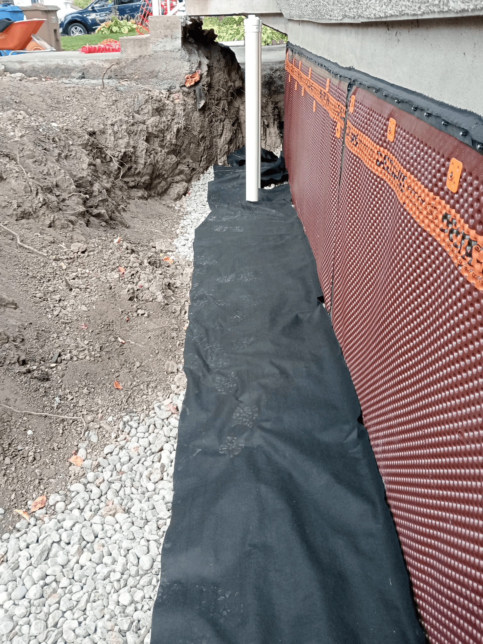 Residential foundation waterproofing work featuring the installation of a brown dimpled drainage membrane on a concrete foundation wall, black geotextile fabric covering a crushed stone bed and an excavation trench, illustrating foundation protection against moisture and water infiltration