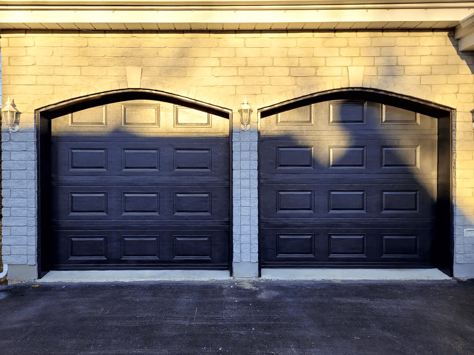 Two modern black garage doors with brick facade and outdoor wall lights.