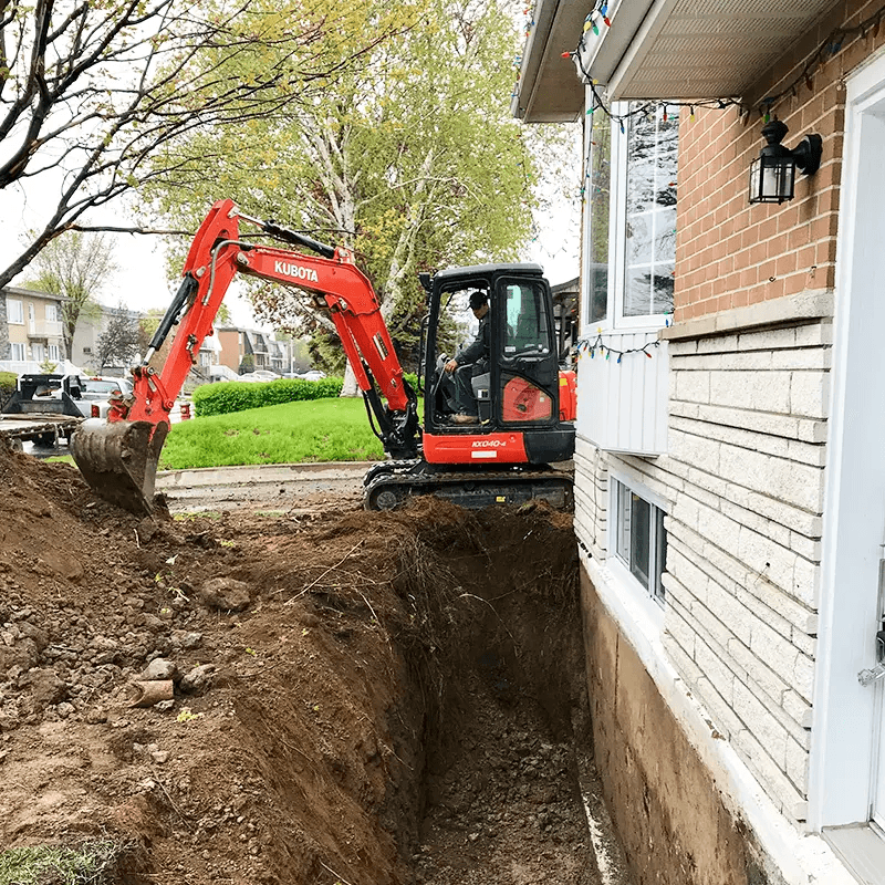 Residential excavation work featuring an orange Kubota mini excavator digging a deep trench along the concrete foundation of a brick house, illustrating site preparation for French drain installation or repair and foundation waterproofing