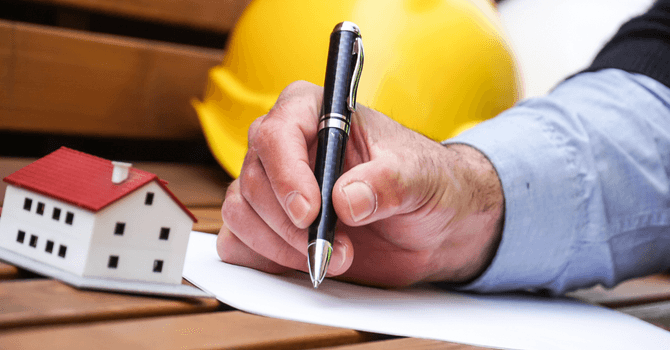 Hand signing a construction document with pen, yellow hard hat and house model on a wooden table.