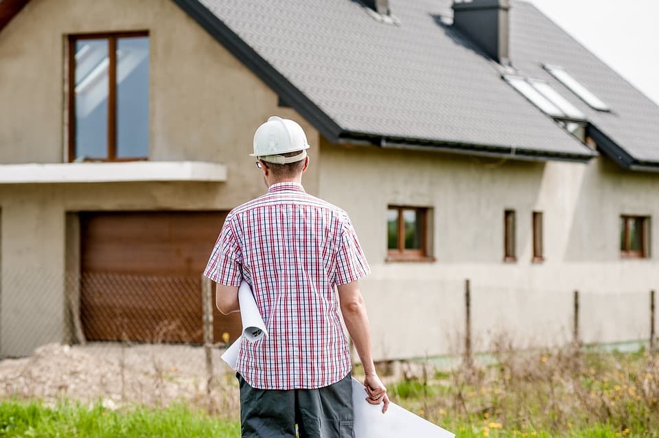 Construction professional wearing a hard hat and holding building plans in front of a residential house, illustrating inspection, planning and management of a home construction or renovation project