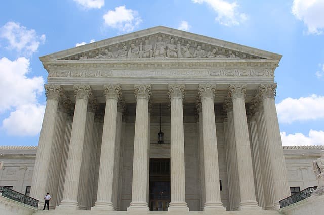 Monumental facade of the United States Supreme Court in Washington DC, featuring imposing white marble Corinthian columns, a sculpted pediment, neoclassical architecture and a grand entrance staircase beneath a blue sky